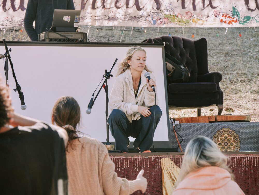 Skylar speaking to a seated audience outdoors with a hand-painted banner